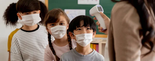 Banner de grupo de estudantes diversos no prédio da escola verificado e escaneado para verificação de temperatura. Alunos elementares estão usando uma máscara facial e se alinham antes de entrar em sala de aula — Fotografia de Stock