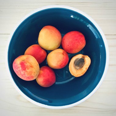 Soluk düz ahşap masa üzerinde koyu mavi bir kapta taze kayısı