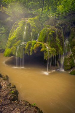 Manzaralı bir manzarada güzel bir şelale, yosunlarla kaplı büyük bir uçurum, arkasında bir köprü ve atmosferinde sis olan bazı kayalar olan bir nehre dökülüyor.