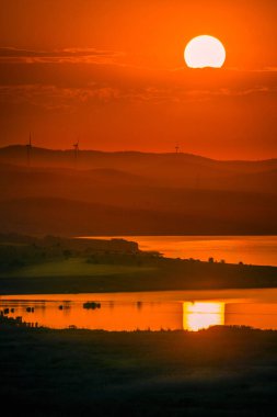 Tepelere yerleştirilen Eolian rüzgar türbinleri günbatımında ekolojik yeşil elektrik üretirken ön planda bir göl ve canlı zengin renkler ve batan güneş.
