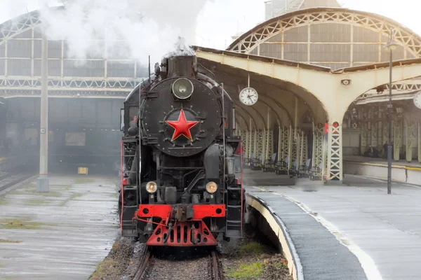 Retro steam train departs from the railway station with the clock on the platform