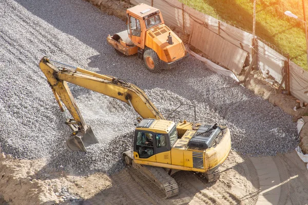黄色挖掘机在瓦砾作业中铺路, 旁边是一个沥青堆垛机. — 图库照片