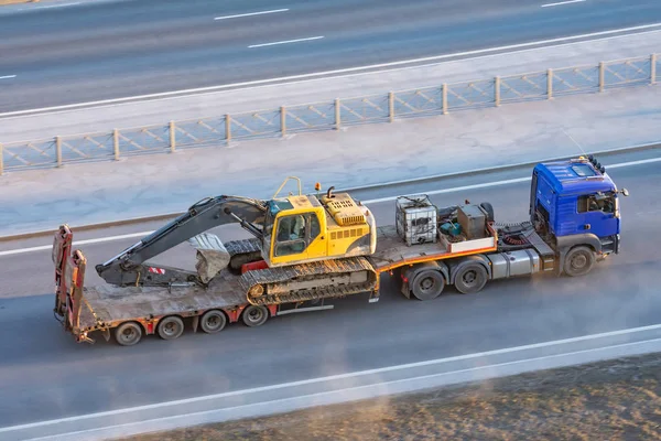 Camión con una plataforma de remolque largo para el transporte de maquinaria pesada, excavadora de orugas cargada con cubo. Transporte por carretera . — Foto de Stock