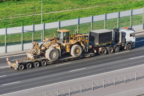 Camión con una plataforma de remolque largo para el transporte de maquinaria pesada, tractor grande cargado con cubo. Transporte por carretera . — Foto de Stock