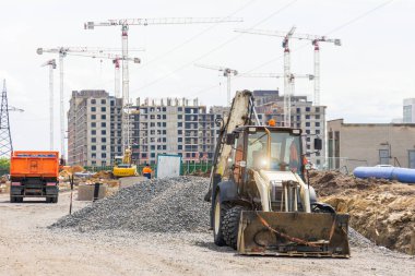 Elinde kova olan traktör, vinç ve çok katlı binaların inşa edildiği bir inşaat sahasında duruyor.