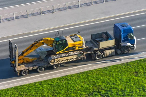 全市公路上装有长挂车月台的运输卡车上的重黄色挖掘机 — 图库照片