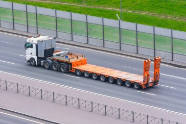 Karavanı ve boş turuncu bir platformu olan bir kamyon şehirde otoyolda ilerliyor.