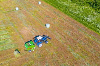 Bir tarım traktörü tarımsal kullanım için biçilmiş çimenleri toplar ve saman balyalarını plastik bir alanda, havadan bakıldığında