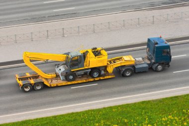Yeni, ağır, sarı kazıcı, uzun, taşıma kamyonunda patlayıcı kovası. Kentteki otoyolda lastik tekerlekler, uzun karavan platformu var.