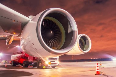 Gece yarısı havaalanında park etmiş büyük bir uçağın kanadı altında iki motor, yanında bir yakıt kamyonu var.