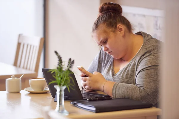 Adolescente con sobrepeso sentada en un café escribiendo mensajes con su teléfono Imágenes De Stock Sin Royalties Gratis