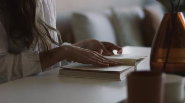 Evde kağıt kitap okuyan bir kadın. İç mekan. El kamerası 4K. Masanın kenarında oturan kız sayfaları çeviriyor. Doğal ışık. Edebiyat eğitimi entellektüel miras aracı alternatifi