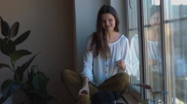 Mutlu kız pencere ev iç yaşam tarzı tarafından kapalı sarı fincan tutarak çay kahve içme. 4k Gülümseyen genç kadın pencere kafede oturan zevk zaman şarkı. Boş zaman kadın güzellik