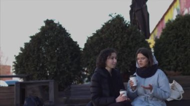 Parkın ön görünümünde dışarıdaki bankta otururken iyi vakit geçiren kadın arkadaşların 4k portresi. Birbirine yakın oturan iki güzel kadın kahve çayı içiyor. Kız arkadaş dostluk