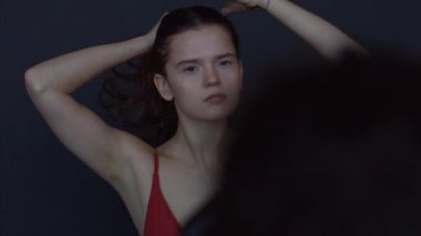 Kırmızı elbiseli genç kadın sahne arkasında fotoshoot 4k pozu veriyor. Erkek eller, arka plandaki siyah stüdyoda saçlarını okşayan kızın fotoğrafını çekiyor. Moda güzellik endüstrisi yüz kozmetiği