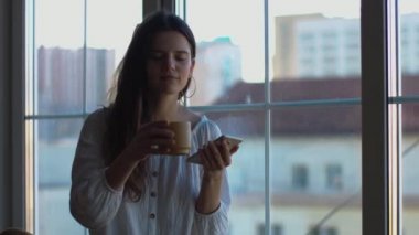 Cep telefonu tutan çekici kız evde doğal ışık sabah kahve çayı içme. Genç kadın pencere içinde 4k oturan cep akıllı telefon yazarak. mutlu modern yaşam tarzı gadget