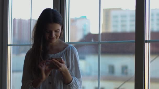 ウィンドウ都市の背景によって屋内に立って携帯電話でオンラインでチャット幸せな女性 ルームフロントビューでスマートフォンのタイピングメッセージを持つ若い女性の4Kビデオショット 人のコミュニケーション — ストック動画