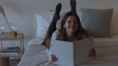Mutlu kız yatak odası önünde 4k el kamera yatan kağıt kitap okuma. Neşeli bayan evde dergi ile zaman zevk kameraya bakarak. Gevşeme sağlık öz bakım kadın güzellik