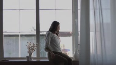 Genç bayan pencereden dışarı bakıyor. Arka planda 4k doğal ışık. Pencere kenarında tek başına oturan düşünceli bir kız el kamerası. Boş zaman yalnızlığı problem çözme