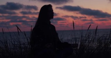 Şafak söktüğünde deniz de tek başına oturan meditasyon kadınınSiyah silueti. Yaz tatili seyahat el cihazı yavaş hareket okyanus kıyısında rahatlatıcı tanınmayan genç kadın