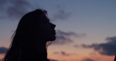 Yaz akşamı yavaş çekimin tadını çıkarırken gökyüzüne bakan uzun saçlı kadının karanlık profili. Gün batımı arka planda tek başına rahatlatıcı siluetli kız yan portre. Meditasyon sağlıklı yaşam stresüstesinden