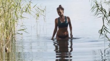 Mayolu kız, açık havada ağır çekimde tek başına banyo yaptıktan sonra nehir suyundan çıkıyor. Cilt manzaralı portrede ıslak damlalarla havuzdan çıkan seksi genç kadın. Yaz tatili seyahati