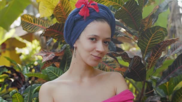 Close Up Portret pięknej młodej dziewczyny w turban na tle natury. Urocza, zmysłowa kobieta patrząca w kamerę i flirtująca na tle dżungli. Powolny film wideo z turystycznej dziewczyny z kwiatem — Wideo stockowe