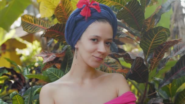 Primer plano retrato de hermosa joven en turbante en fondo de la naturaleza. Encantadora mujer sensual mirando a la cámara y coqueteando en el fondo de la selva. Imágenes de cámara lenta de chica turística con flor — Vídeos de Stock