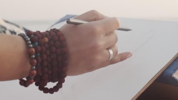 Vista de cerca de la mujer haciendo boceto al aire libre en el fondo de la playa del mar dispositivo de mano. Artista sin rostro dibujando con lápiz sentado en la playa solo en cámara lenta. Artesanía hobby verano — Vídeos de Stock