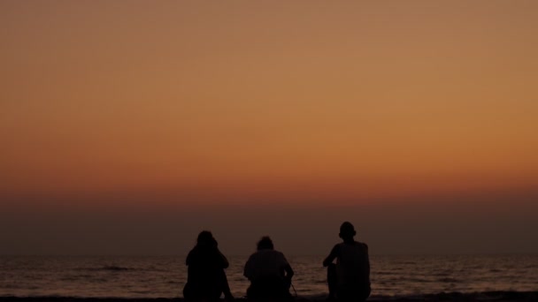 Черные силуэты неузнаваемых туристов, сидящих на пляже и смотрящих на закат океана. Три человека отдыхают на берегу моря вместе в оранжевом вечернем замедленном движении. Счастье в отпуске — стоковое видео