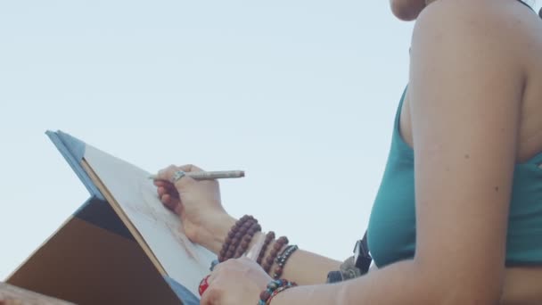 Nízký úhel pohledu dívky kreslení náčrtek na papírové podložce venku na modrém pozadí oblohy. Umělec bez tváře pracuje na obrázku na letní teplý den makro detail. tvůrčí proces workshopu — Stock video