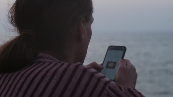 De cerca sobre la vista del hombro del hombre desplazándose noticias sobre el fondo desenfocado del mar. Hombre sin rostro sosteniendo smartphone leyendo noticias de última hora al aire libre cámara lenta copiar el espacio de texto. Concepto de información — Vídeo de stock