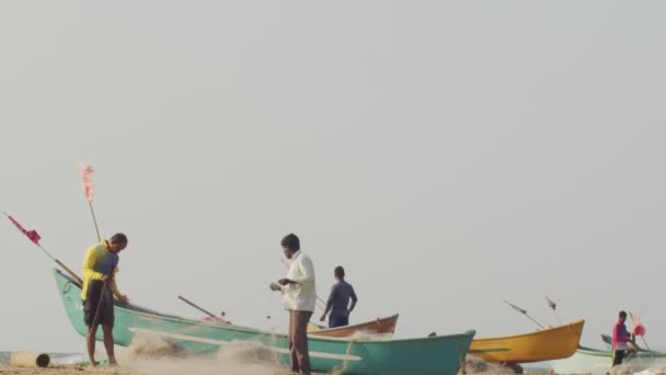 GOKARNA, INDIEN-KRUARI 02,2020: Indiska fiskare kontrollerar fiskenät stående vid sandstrand av färgglada båtar kopiera utrymme text. Autentiska människor förbereder sig för att segla på solig sommardag bakgrund — Stockvideo