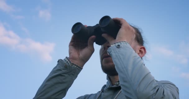 青い空の背景にスローモーションでフィールドガラスを探して旅行者の低角度ビュー。双眼鏡で地平線を探検する真剣な男屋外のハンドヘルドデバイス。夏休み旅行発見 — ストック動画