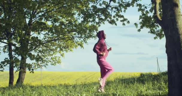 Vista lateral de la atleta femenina en ropa deportiva rosa corriendo en el lugar al aire libre en la naturaleza salvaje. Mujer joven entrenando trotando sola en cámara lenta de aire fresco. Estilo de vida deportivo concepto de cuidado corporal de salud — Vídeos de Stock