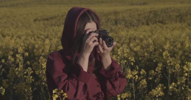 Outdoor-Porträt eines Mädchens, das die Natur auf einem Handheld-Gerät mit Vintage-Kamera fotografiert. Junge Fotografin fotografiert im Freien gelbe Feld Hintergrund Zeitlupe. Sommerreise zum Erinnerungsglück — Stockvideo