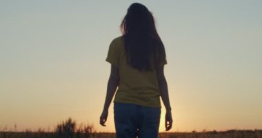 Tanımlanamayan bir kızın doğanın arka planında yavaş çekimde gün batımına gidişinin arka görüntüsü. Uzun saçlı kadın, altın güneş ışığının sığ ışıklarıyla tarlada tek başına yürüyor. Mutlu yaşam tarzı konsepti
