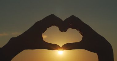 Turuncu günbatımı arka planında kalp işareti siluetini tutan kadın ellerini kapat. Aşkın sembolünü gösteren meçhul kadın güneşin yavaş çekimde çekilmesiyle arka planda kaldı. Umut bakım konseptine güven