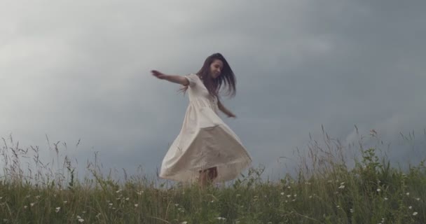 Mladá žena v ženských šatech se před deštěm obrací venku na zeleném poli. Šťastná dívka tančí rotující sám na venkově zpomalit kopírování textu prostoru. Svoboda štěstí letní cestování — Stock video