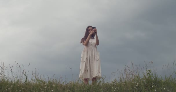 Low angle view of girl in vintage dress making mobile photo of nature outdoors copy text space. Young woman photographing on smartphone standing in field countryside handheld device. Travel technology — Stock Video