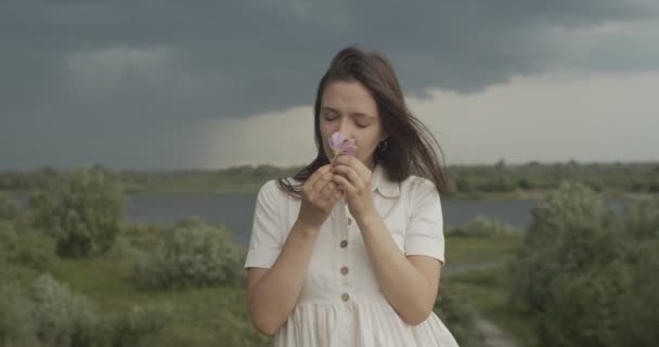 Elegant girl in white dress holding wild flower outdoors on stormy landscape background. Long-haired woman breathing rainy air on summer windy day slow motion. Religion happiness trust hope concept — Stock Video