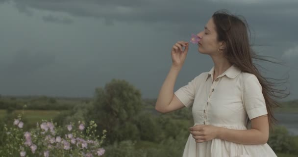 Side portré lány szagú vad virág a szabadban viharos égbolt ország háttér. Gyönyörű nő élvezi a természet gazdaság növény lassított felvétel szöveges hely. Szépségápolási termékek környezete — Stock videók