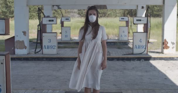 Serious girl wearing anti-virus mask on empty gas-station slow motion. Portrait of long-haired woman standing alone during covid quarantine outdoors. Pandemic infection disease concept — Stock Video