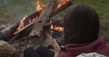 Ateşin yanında oturan tanınmayan bir kız. Yakın plan akıllı telefon kullanıyor. Yüzü olmayan esmer kadın, dışarıda ateşin yanında oturan sandviçin fotoğrafını çekiyor. Turist Doğada Zaman Geçiren Şenlik Ateşi 'nin Yakınında Isınıyor