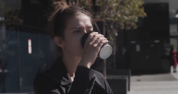 Mladá žena pije kávu z jednorázové pohár venku na slunném městském pozadí. Přírodní dívka drží čaj nápoj na ulici těší slunce zpomalení pohybu. Energie kofein kultura potravin životní styl — Stock video
