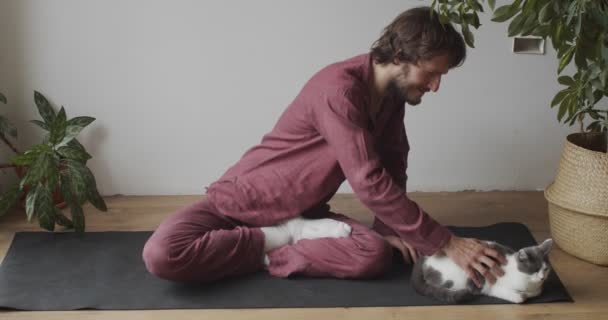 Чоловік сидить на йога килимок схрещений погладжуючий пухнастий кіт в приміщенні природного світла повільний рух. Йоги майстер розслабляється перед тренуванням медитації. Спортивна охорона здоров'я активний спосіб життя. Коханці тварин — стокове відео