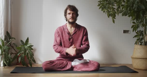 Interior retrato de homem barbudo segurando Namaste mãos saudando câmera lenta. Mestre de iogue caucasiano sentado de pernas cruzadas no tapete com as mãos unidas luz natural. Meditação estresse superar a prática de ioga — Vídeo de Stock