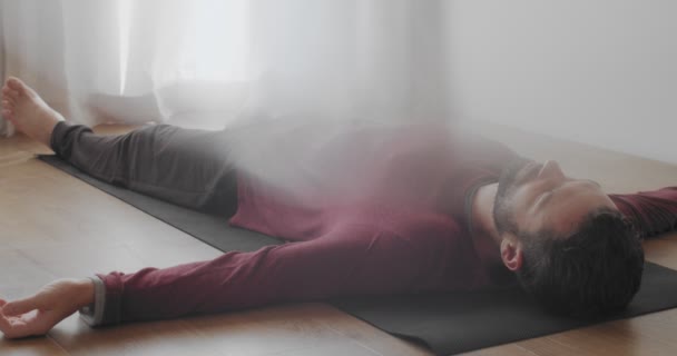 Vue à angle bas de l'homme se relaxant dans shavasana asana intérieur lumière naturelle du matin. Barbu instructeur caucasien couché sur tapis de yoga après la pratique au ralenti. relaxation méditation stress surmonter — Video