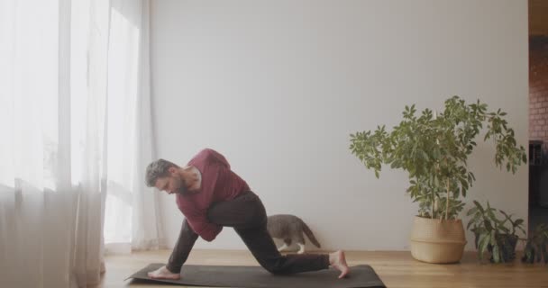 Uomo bianco praticare yoga all'interno su sfondo bianco parete luce naturale soleggiata. Yogi barbuto in piedi in equilibrio in posa su mat slow motion. Flessibilità allungamento lavoro del corpo. Abitudini di vita sane — Video Stock