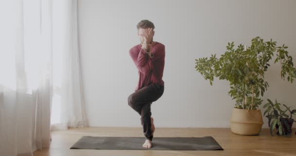 白地のスローモーションで屋内にGarudasana asanaに立つ若いトレーナー。自宅で一人でハタヨガを練習しているヨギ男の肖像画。集中瞑想｜健康医療 — ストック動画
